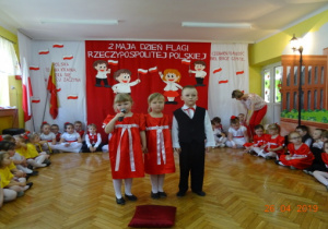 2 dziewczynki w czerwonych sukienkach i chłopiec ubrany na galowo recytują wiersz.